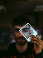 A man with dark hair and beard is holding a Polaroid photo in front of his face. The photo captures a close-up of his eye, adding a sense of depth and intrigue. The background is blurred, drawing focus to the eye and facial features in the Polaroid.