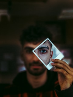 A man with dark hair and beard is holding a Polaroid photo in front of his face. The photo captures a close-up of his eye, adding a sense of depth and intrigue. The background is blurred, drawing focus to the eye and facial features in the Polaroid.