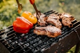 Several pieces of grilled meat are sizzling on a black barbecue grill, accompanied by red and yellow bell peppers. The grill is situated outdoors with a blurred background of green foliage, indicating a sunny and fresh atmosphere.