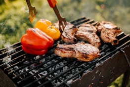 Several pieces of grilled meat are sizzling on a black barbecue grill, accompanied by red and yellow bell peppers. The grill is situated outdoors with a blurred background of green foliage, indicating a sunny and fresh atmosphere.