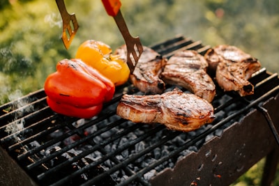 Several pieces of grilled meat are sizzling on a black barbecue grill, accompanied by red and yellow bell peppers. The grill is situated outdoors with a blurred background of green foliage, indicating a sunny and fresh atmosphere.