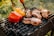 Several pieces of grilled meat are sizzling on a black barbecue grill, accompanied by red and yellow bell peppers. The grill is situated outdoors with a blurred background of green foliage, indicating a sunny and fresh atmosphere.