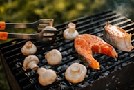 Grill with two fish steaks and several mushrooms being cooked. There is a pair of tongs with wooden handles resting on the grill.
