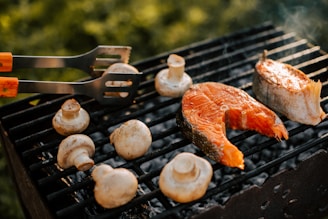 Grill with two fish steaks and several mushrooms being cooked. There is a pair of tongs with wooden handles resting on the grill.