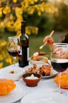A table set outdoors with a variety of dishes including pumpkin, wine, olives, and a serving of meat. Two glasses of red wine are prominently placed alongside a bottle. The hand of a person is using a fork to pick up a piece of food, creating a cozy and inviting dining atmosphere.