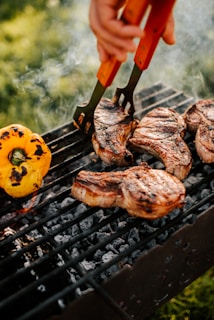 Juicy steaks grilling over charcoal with a vibrant yellow bell pepper next to them. A hand uses tongs to flip the meat, creating a mouthwatering, smoky scene on the barbecue. The background is a blur of greenery, suggesting an outdoor setting.