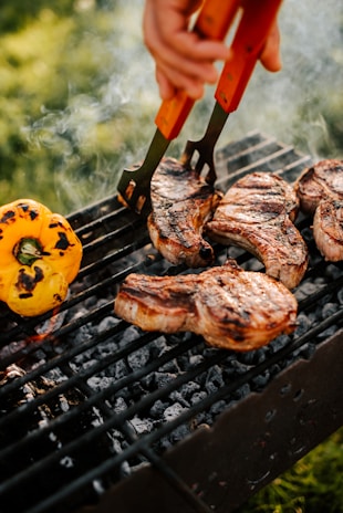 Juicy steaks grilling over charcoal with a vibrant yellow bell pepper next to them. A hand uses tongs to flip the meat, creating a mouthwatering, smoky scene on the barbecue. The background is a blur of greenery, suggesting an outdoor setting.
