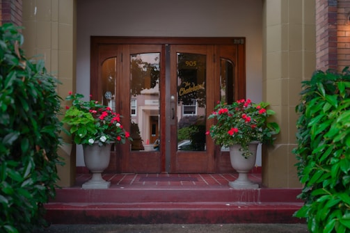 A wooden double door entrance is flanked by large potted plants with vibrant red and white flowers on a red-brick stoop. The pots are symmetrical, creating a welcoming and elegant facade under the name 'The Chatelaine' with the number 905 above the doors. Green foliage surrounds the entrance, adding lushness.