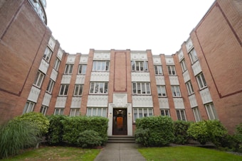 A multi-story brick apartment building with white decorative accents around the windows and door. The architecture features intricate patterns and neatly trimmed bushes line the entrance. The building's facade is symmetrical with a central entrance, and there is greenery in the foreground.