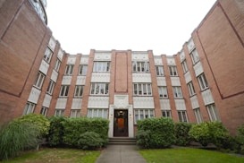 A multi-story brick apartment building with white decorative accents around the windows and door. The architecture features intricate patterns and neatly trimmed bushes line the entrance. The building's facade is symmetrical with a central entrance, and there is greenery in the foreground.