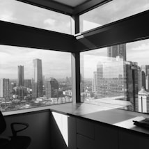 A view from inside an office with large windows showcasing a city skyline filled with modern skyscrapers. The image is in black and white, giving a timeless and classic feel. The office appears empty, with a chair and a desk positioned in the foreground, highlighting the stark contrast between the interior and the vast urban landscape outside.