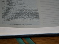 A close-up of an open book with text primarily discussing a prophecy or a statement from the Sovereign Lord. The text is in English, possibly from a religious scripture, and has a structured layout on the page. The background includes a wooden surface and a green ribbon marker.