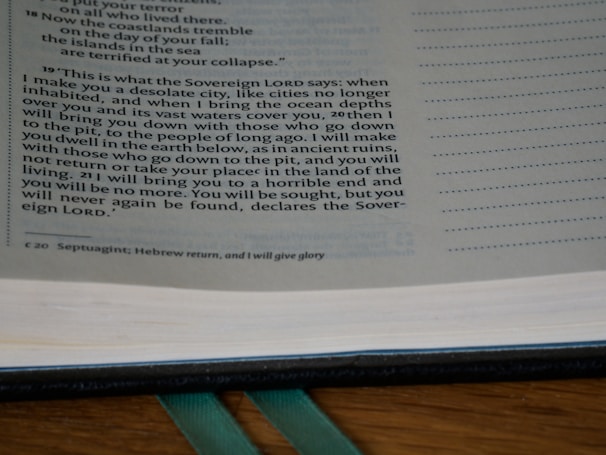 A close-up of an open book with text primarily discussing a prophecy or a statement from the Sovereign Lord. The text is in English, possibly from a religious scripture, and has a structured layout on the page. The background includes a wooden surface and a green ribbon marker.