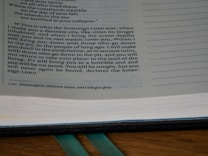 A close-up of an open book with text primarily discussing a prophecy or a statement from the Sovereign Lord. The text is in English, possibly from a religious scripture, and has a structured layout on the page. The background includes a wooden surface and a green ribbon marker.