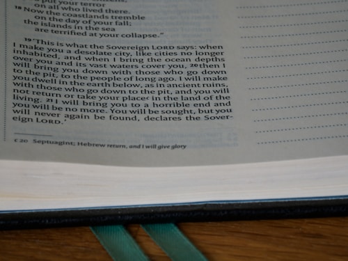A close-up of an open book with text primarily discussing a prophecy or a statement from the Sovereign Lord. The text is in English, possibly from a religious scripture, and has a structured layout on the page. The background includes a wooden surface and a green ribbon marker.