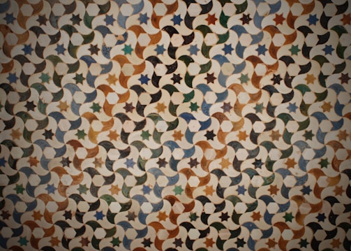 An intricate geometric pattern consisting of repeating stars and crescent shapes. The design features a combination of earthy tones with variations of green, brown, and blue. Each element connects seamlessly, creating a continuous visual flow that gives a sense of depth and rhythm.
