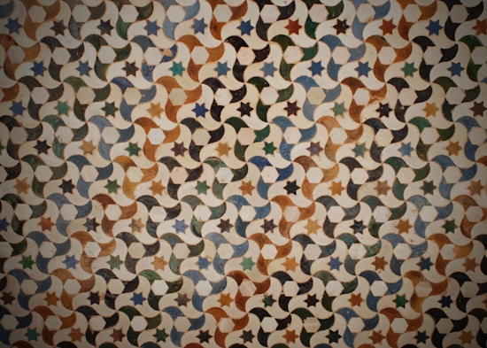An intricate geometric pattern consisting of repeating stars and crescent shapes. The design features a combination of earthy tones with variations of green, brown, and blue. Each element connects seamlessly, creating a continuous visual flow that gives a sense of depth and rhythm.