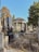 A historical cemetery with various gravestones and monuments, featuring an ancient pavilion structure with columns in the background. The gravestones are made of stone and some have Hebrew inscriptions. Vegetation is sparse, with overgrown weeds and trees surrounding the area. The overall scene strikes a balance between nature and man-made structures.