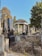 A historical cemetery with various gravestones and monuments, featuring an ancient pavilion structure with columns in the background. The gravestones are made of stone and some have Hebrew inscriptions. Vegetation is sparse, with overgrown weeds and trees surrounding the area. The overall scene strikes a balance between nature and man-made structures.