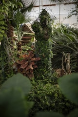 A serene garden scene features a statue covered in lush greenery, surrounded by dense foliage. Various plants, including a palm tree and large leafed vegetation, create a natural, tranquil atmosphere.