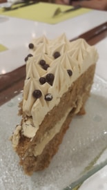 A slice of cake with a beige frosting topped with small chocolate balls is placed on a glass plate. The cake has a layered structure with a light filling and has an overall soft and crumbly texture.