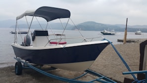 A motorboat with a canopy is positioned on a metal trailer by a sandy beach. The background features a calm body of water with several other boats floating, and distant hills are under an overcast sky.