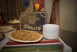 A decorative display featuring a chalkboard sign with 'Viva Mexico' written on it, adorned with an illustrated sombrero. In front of the sign, there is a platter of Mexican food, possibly tacos or quesadillas, arranged neatly. Next to the platter, a stack of white plates is ready for serving. A vibrant striped textile covers the table, and a wire basket with colorful bell peppers is visible in the background.