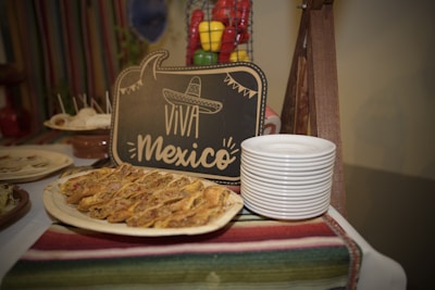 A decorative display featuring a chalkboard sign with 'Viva Mexico' written on it, adorned with an illustrated sombrero. In front of the sign, there is a platter of Mexican food, possibly tacos or quesadillas, arranged neatly. Next to the platter, a stack of white plates is ready for serving. A vibrant striped textile covers the table, and a wire basket with colorful bell peppers is visible in the background.