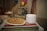 A decorative display featuring a chalkboard sign with 'Viva Mexico' written on it, adorned with an illustrated sombrero. In front of the sign, there is a platter of Mexican food, possibly tacos or quesadillas, arranged neatly. Next to the platter, a stack of white plates is ready for serving. A vibrant striped textile covers the table, and a wire basket with colorful bell peppers is visible in the background.