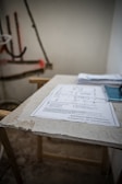 A sheet of paper with technical drawings and text is placed on a rough surface, likely a table. The background is blurry, showing some indistinct objects and a partially visible wall, suggesting a work or construction environment.