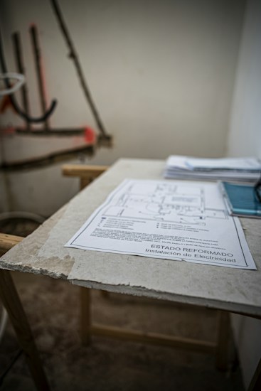 A sheet of paper with technical drawings and text is placed on a rough surface, likely a table. The background is blurry, showing some indistinct objects and a partially visible wall, suggesting a work or construction environment.