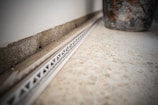 A close-up view of a tile floor with a white plastic spacer or track against the wall. The wall has a rough concrete texture and there is a partially visible, large, decorative pot on the right side. The focus is on the geometric patterns and textures.