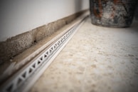 A close-up view of a tile floor with a white plastic spacer or track against the wall. The wall has a rough concrete texture and there is a partially visible, large, decorative pot on the right side. The focus is on the geometric patterns and textures.