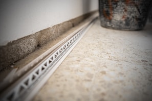 A close-up view of a tile floor with a white plastic spacer or track against the wall. The wall has a rough concrete texture and there is a partially visible, large, decorative pot on the right side. The focus is on the geometric patterns and textures.