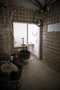 This image depicts an unfinished room under construction. The walls are made of concrete blocks, and there are several construction materials scattered around, including buckets, tools, and cables. A wooden stool is visible near a white door that leads to another area, which appears brighter.