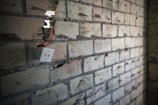A partially destroyed brick wall shows an exposed electrical outlet with visible wiring. The bricks are a mix of gray and beige colors, with cracks and holes. The scene conveys a sense of neglect or incomplete renovation.