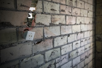 A partially destroyed brick wall shows an exposed electrical outlet with visible wiring. The bricks are a mix of gray and beige colors, with cracks and holes. The scene conveys a sense of neglect or incomplete renovation.