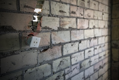 A partially destroyed brick wall shows an exposed electrical outlet with visible wiring. The bricks are a mix of gray and beige colors, with cracks and holes. The scene conveys a sense of neglect or incomplete renovation.