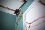 A close-up view of a damaged wall with exposed bricks and visible drywall. There is a metal bracket attached to the wall, possibly used for mounting. The wall features graffiti markings in orange, and there appears to be electrical wiring present.