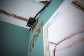A close-up view of a damaged wall with exposed bricks and visible drywall. There is a metal bracket attached to the wall, possibly used for mounting. The wall features graffiti markings in orange, and there appears to be electrical wiring present.