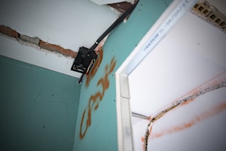 A close-up view of a damaged wall with exposed bricks and visible drywall. There is a metal bracket attached to the wall, possibly used for mounting. The wall features graffiti markings in orange, and there appears to be electrical wiring present.