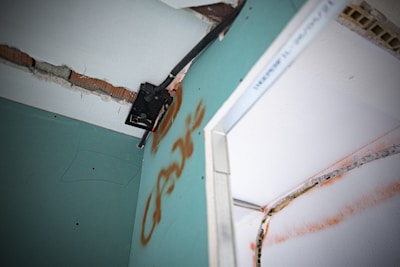 A close-up view of a damaged wall with exposed bricks and visible drywall. There is a metal bracket attached to the wall, possibly used for mounting. The wall features graffiti markings in orange, and there appears to be electrical wiring present.