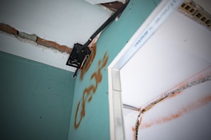 A close-up view of a damaged wall with exposed bricks and visible drywall. There is a metal bracket attached to the wall, possibly used for mounting. The wall features graffiti markings in orange, and there appears to be electrical wiring present.