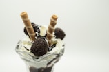 A glass dessert dish filled with vanilla ice cream topped with chocolate syrup, chocolate-covered candies, and two rolled wafer sticks. The presentation is elegant and delicious.