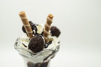 A glass dessert dish filled with vanilla ice cream topped with chocolate syrup, chocolate-covered candies, and two rolled wafer sticks. The presentation is elegant and delicious.