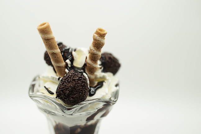 A glass dessert dish filled with vanilla ice cream topped with chocolate syrup, chocolate-covered candies, and two rolled wafer sticks. The presentation is elegant and delicious.