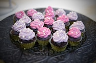 A cheerful baker in a pink apron holding a tray of freshly baked cupcakes with gold frosting.