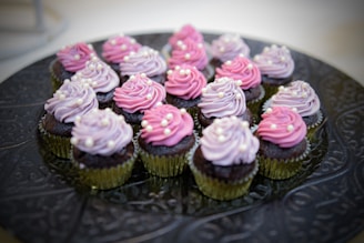 A tray filled with multiple chocolate cupcakes, each topped with swirls of pink and lavender frosting, adorned with small white pearl-like decorations. The cupcakes are placed in golden foil wrappers on an ornate, dark-colored platter.