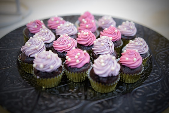 A tray filled with multiple chocolate cupcakes, each topped with swirls of pink and lavender frosting, adorned with small white pearl-like decorations. The cupcakes are placed in golden foil wrappers on an ornate, dark-colored platter.