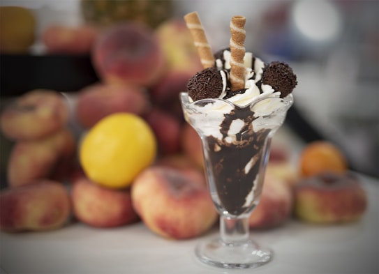 A dessert glass filled with layers of chocolate syrup and whipped cream, topped with two chocolate truffles and two wafer sticks. The background is blurred with various fruits including peaches and a lemon.